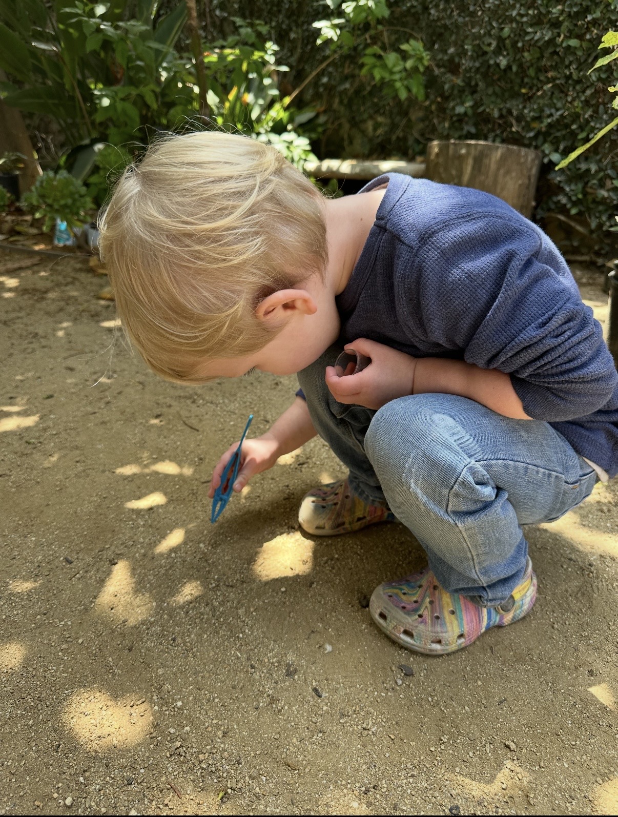 Toddler crouching outside in the garden exploring the ground with a toy tool
