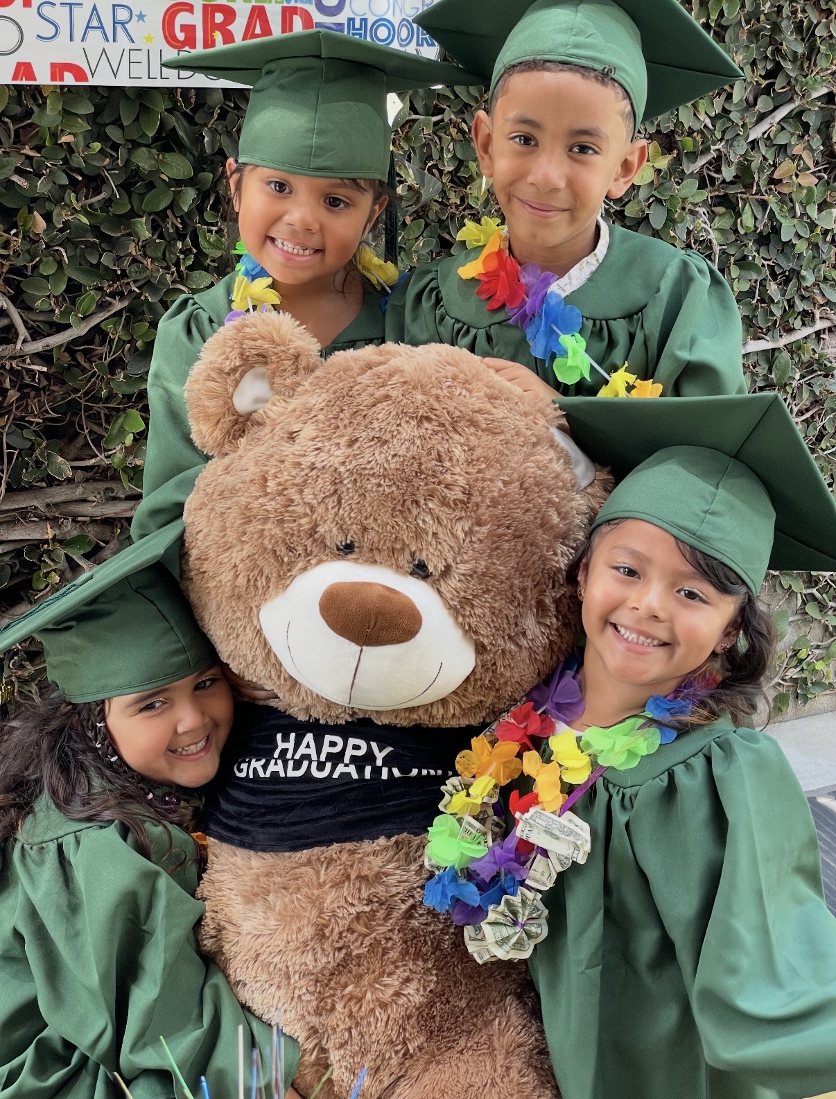Four preschool graduates in green caps and gowns hugging a giant teddy bear