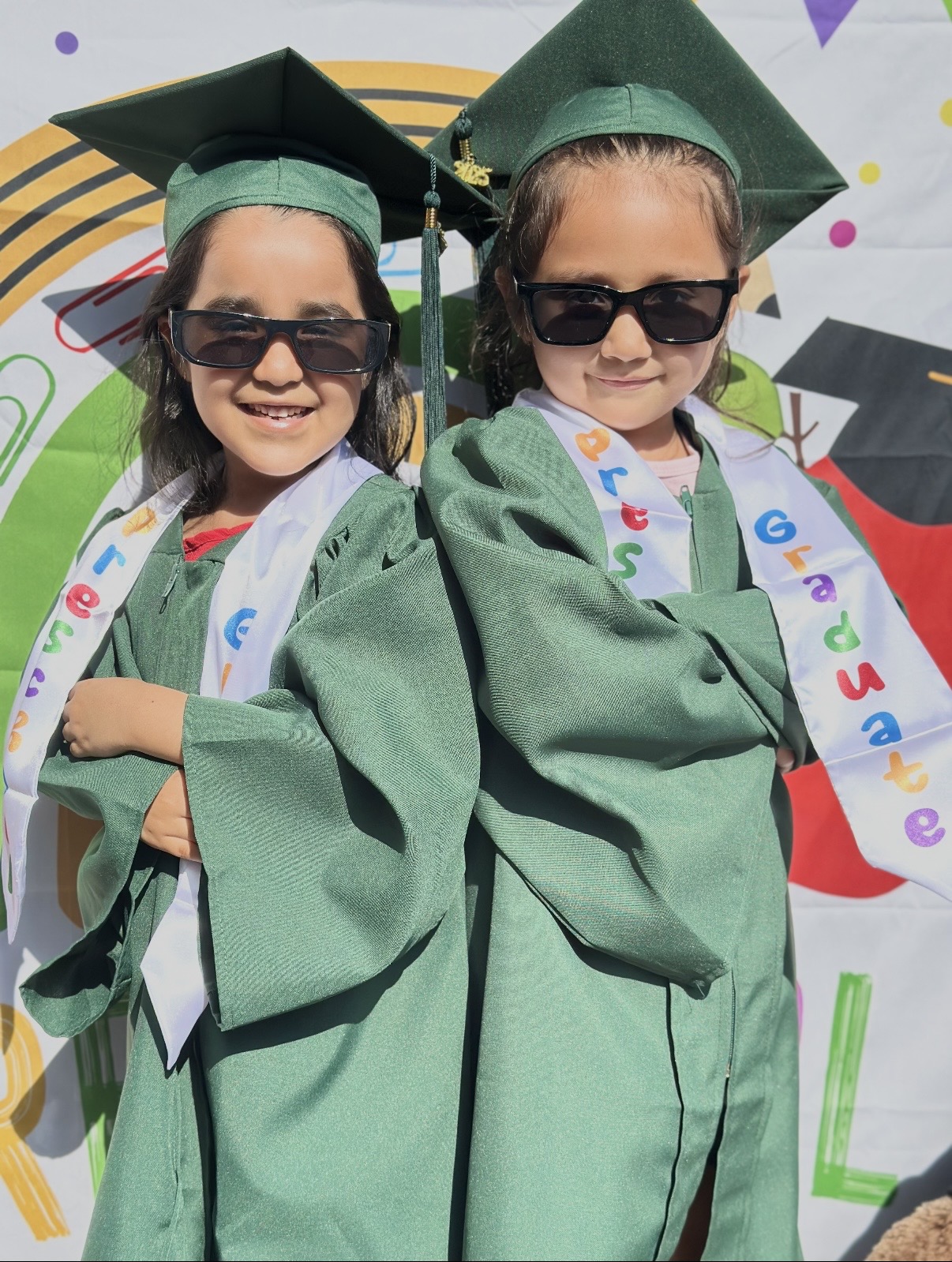 Two preschool graduates in green caps and gowns wearing sunglasses